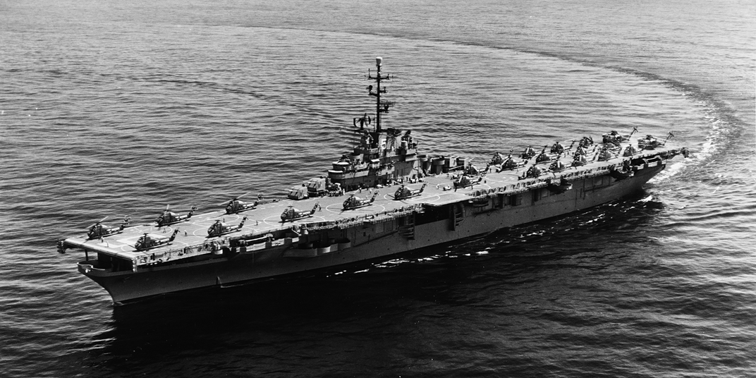 A black-and-white photo of a small aircraft carrier making a sweeping turn on a calm sea.